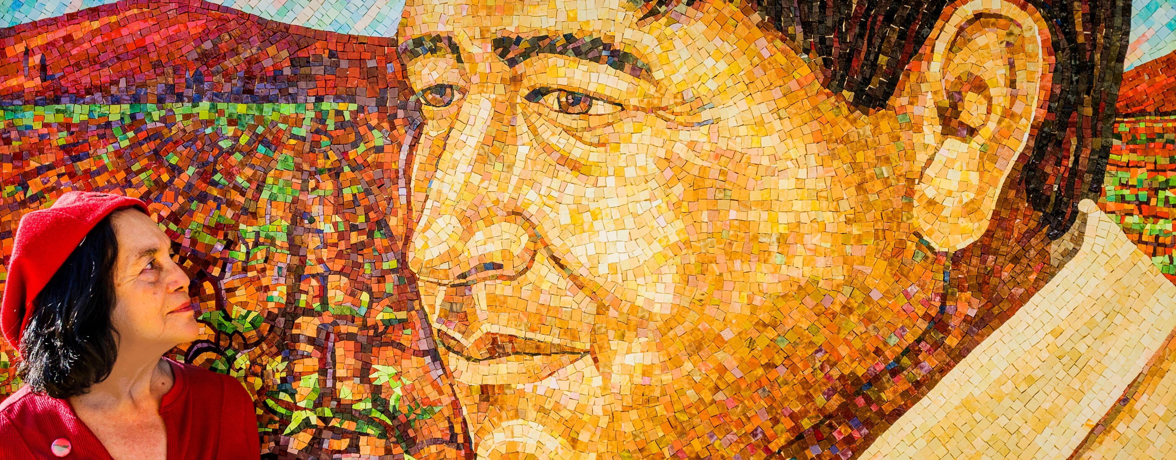 Dolores Huerta in a red hat and top lovingly looks at the colorful mosaic of César Chávez.