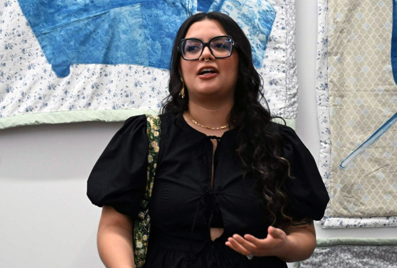 A woman with glasses stands next to a collection of blue and white quilts hung on the wall, emphasizing the vibrant colors and patterns.