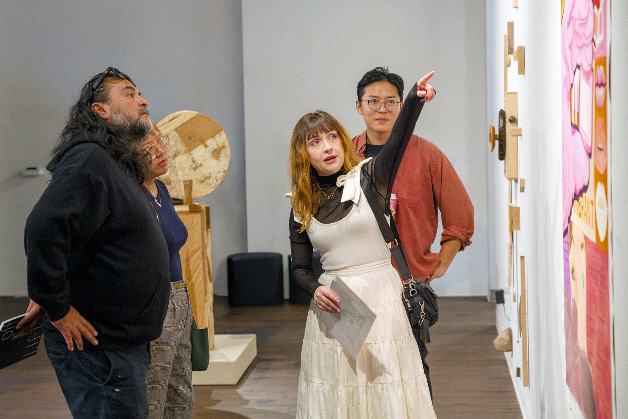 An artist points at her painting in a gallery, inviting viewers to observe the artwork closely.