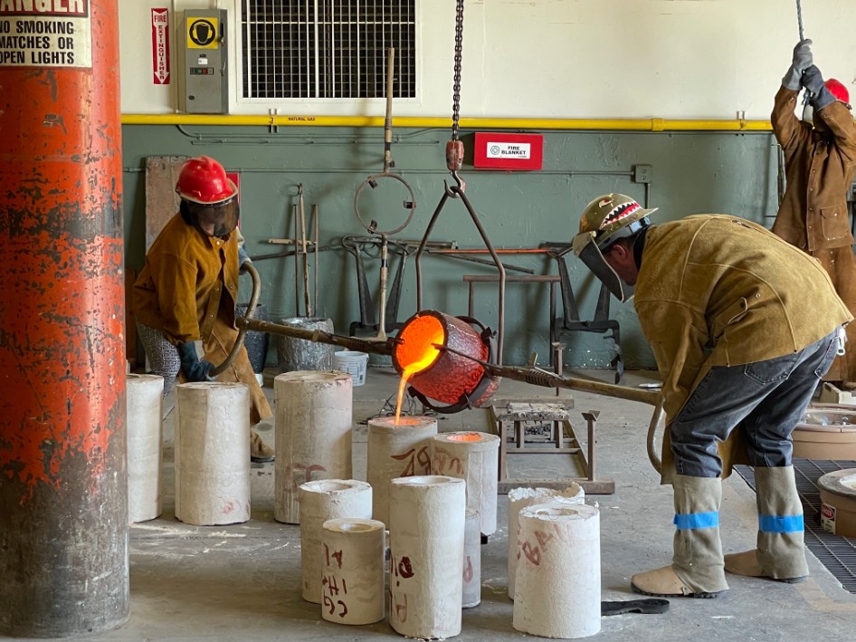 Two students in hard hats and safety glasses help pour molten bronze into molds in a foundry