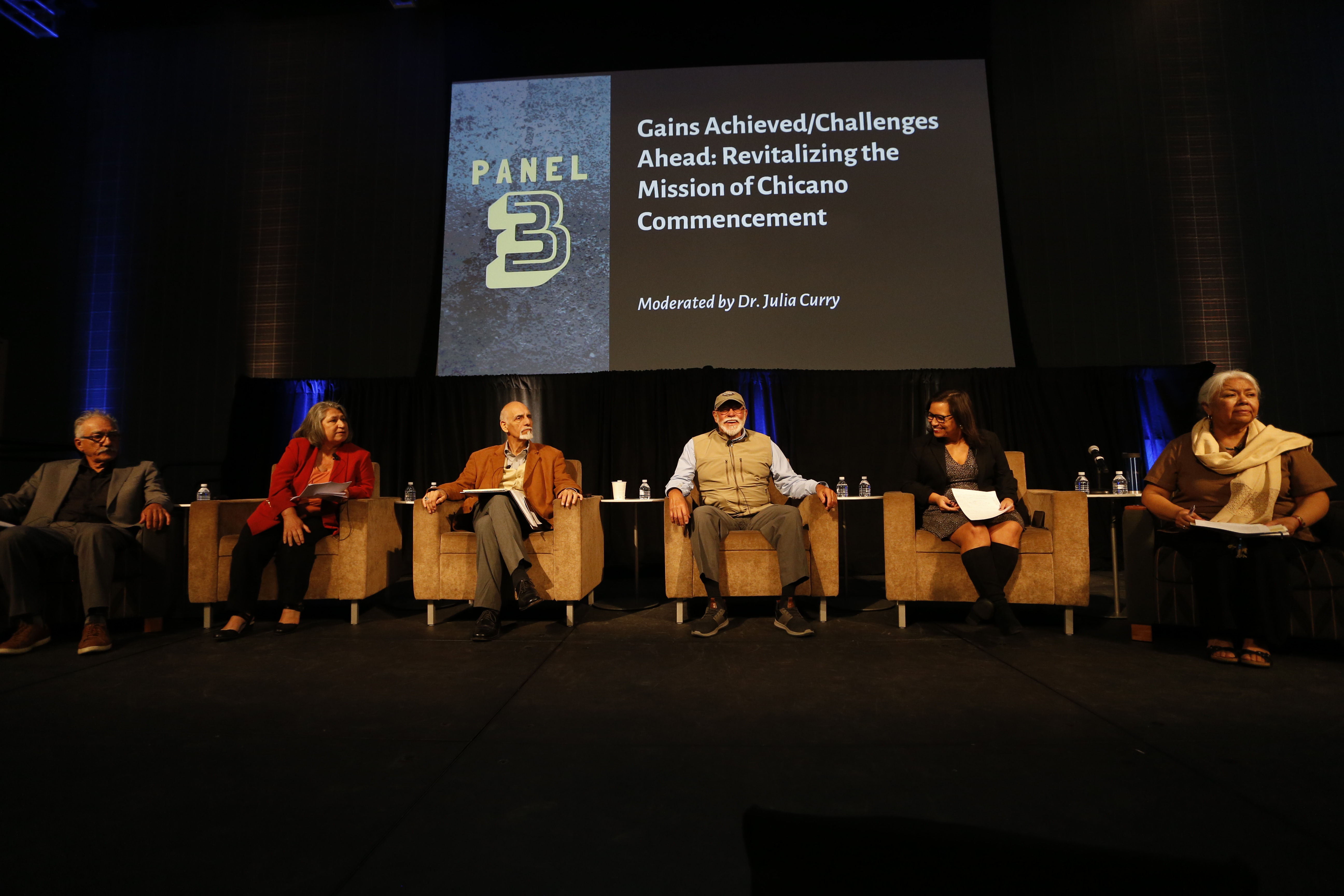 A group of older and younger alumni sit as a panel on stage in front of a screen.