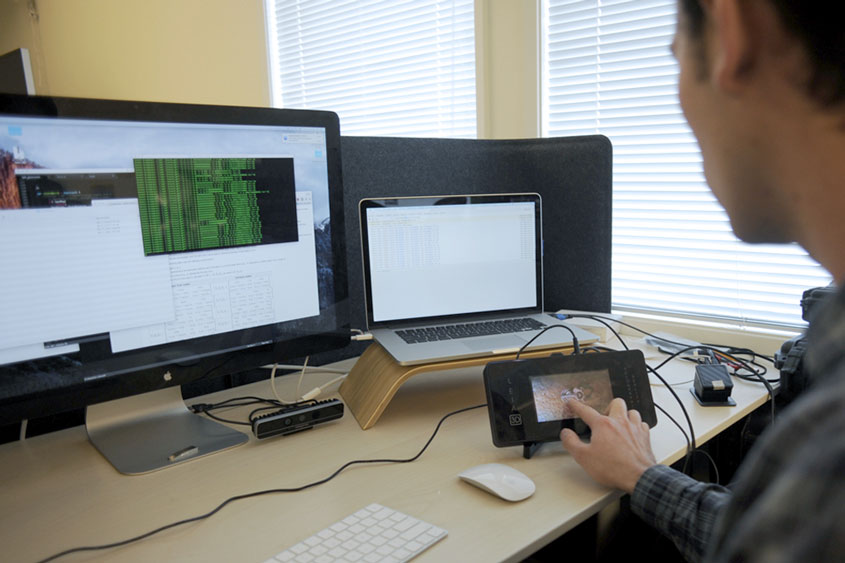 Male student touching a handheld device that is plugged into a computer.