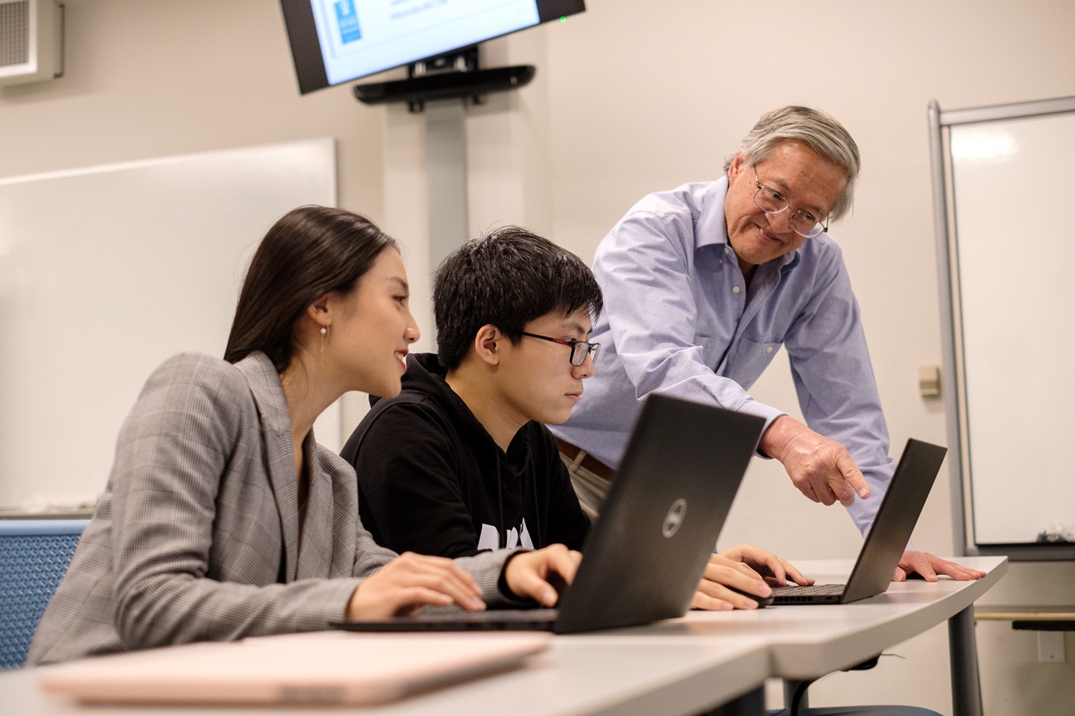 Two students sit at a desk in front of a computer with a professor teaching them at SJSU. 