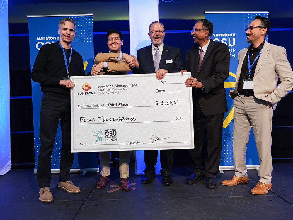 A college-age startup founder poses onstage with two sponsors, two hosts, and a giant check for $5,000