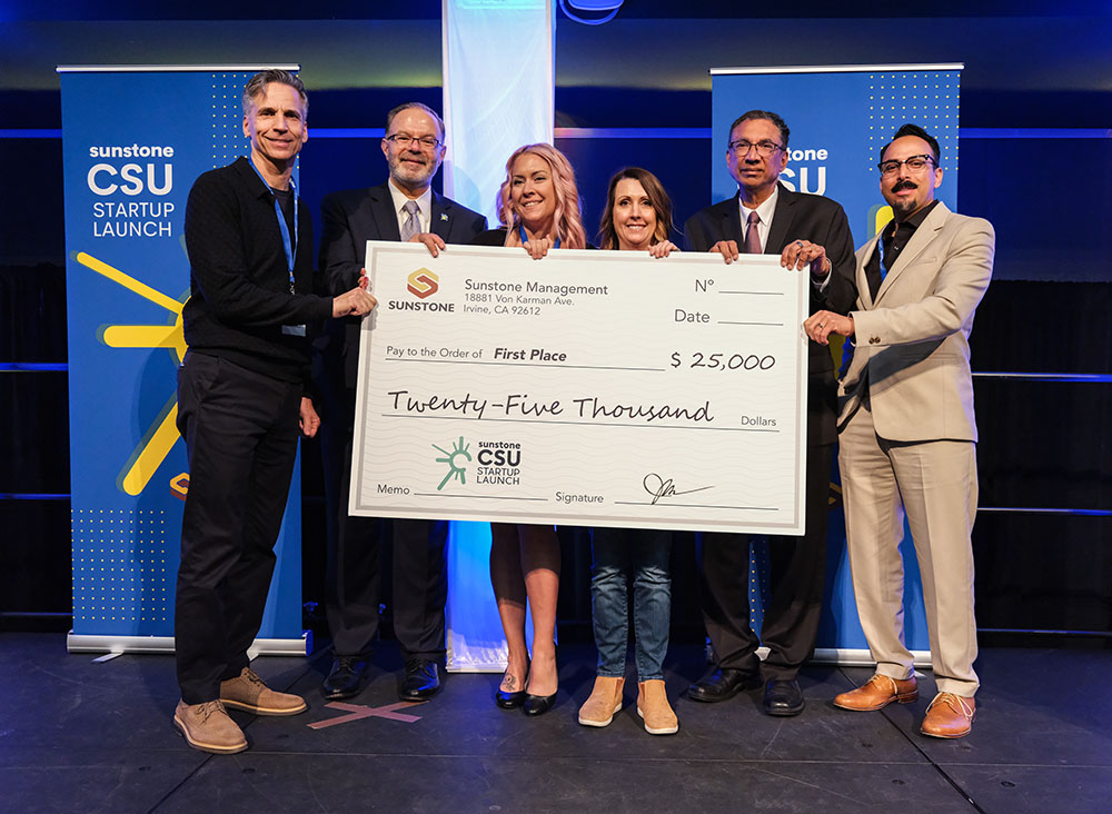 Two female, college-age startup founders pose onstage with two sponsors, two hosts, and a giant check for $25,000