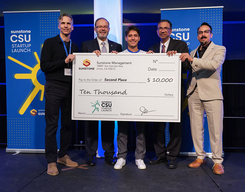 A male, college-age startup founder poses onstage with two sponsors, two hosts, and a giant check for $10,000