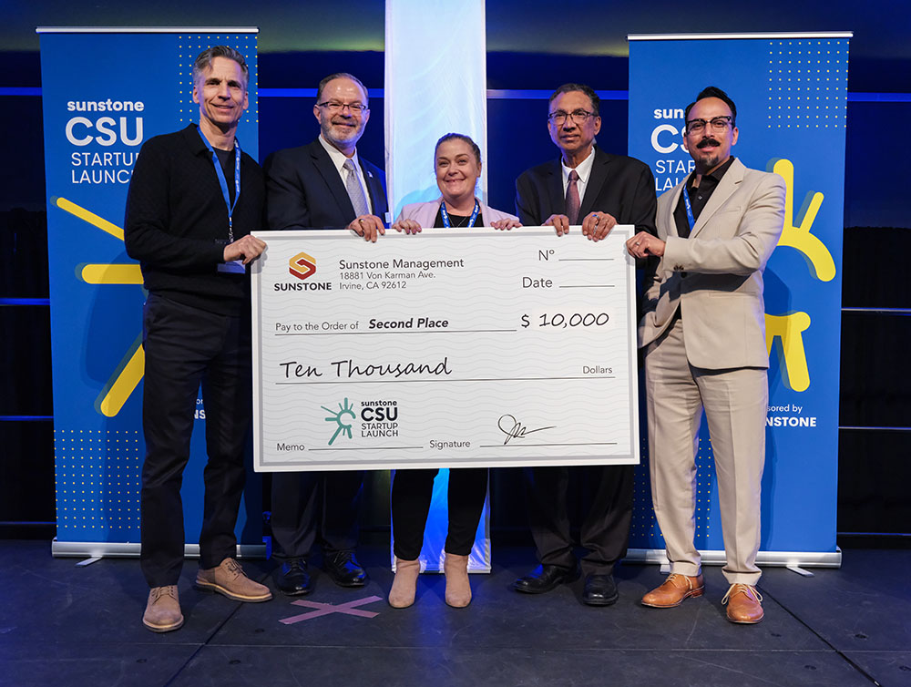 A female, college-age startup founder poses onstage with two sponsors, two hosts, and a giant check for $10,000