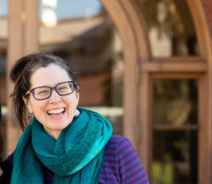 A white woman with dark brown hair and black glasses, wearing a blue and purple striped shirt