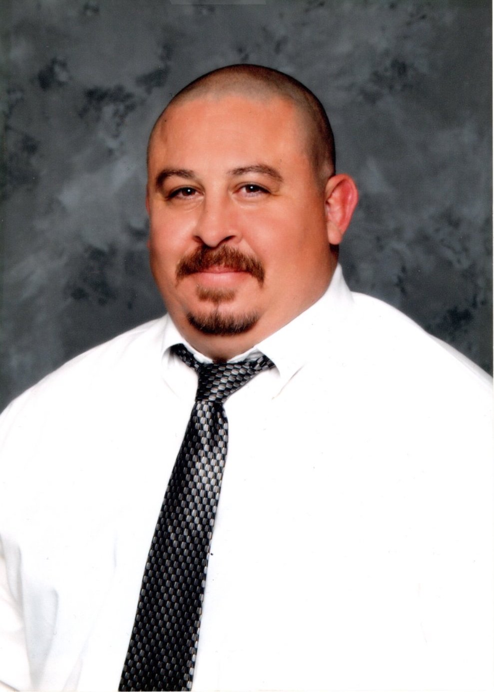 Hispanic male with buzzed, brown hair and a brown moustache, wearing a white button-down and black tie, smiling in front of a black background.