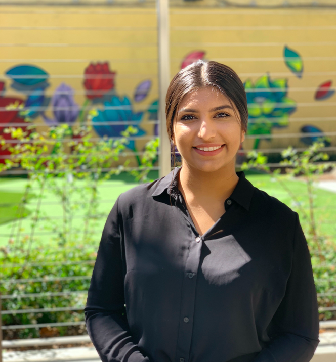 Middle Eastern woman with black hair in a ponytail, wearing a black button down shirt, smiling in front of a painted elementary school wall. 