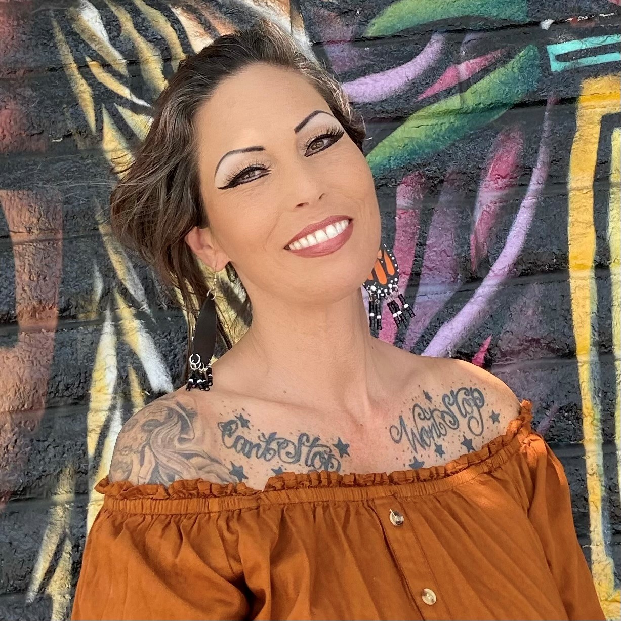 Hispanic woman with tattoos near her shouler, with medium length brown hair, wearing an orange shirt, smiling in front of grafitied wall. 