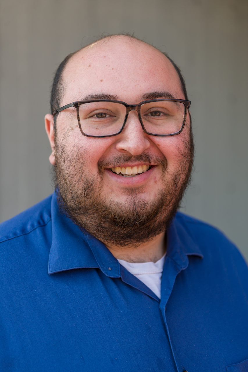 Male in a blue, collared shirt, wearing glasses, smiling in front of a gray background.