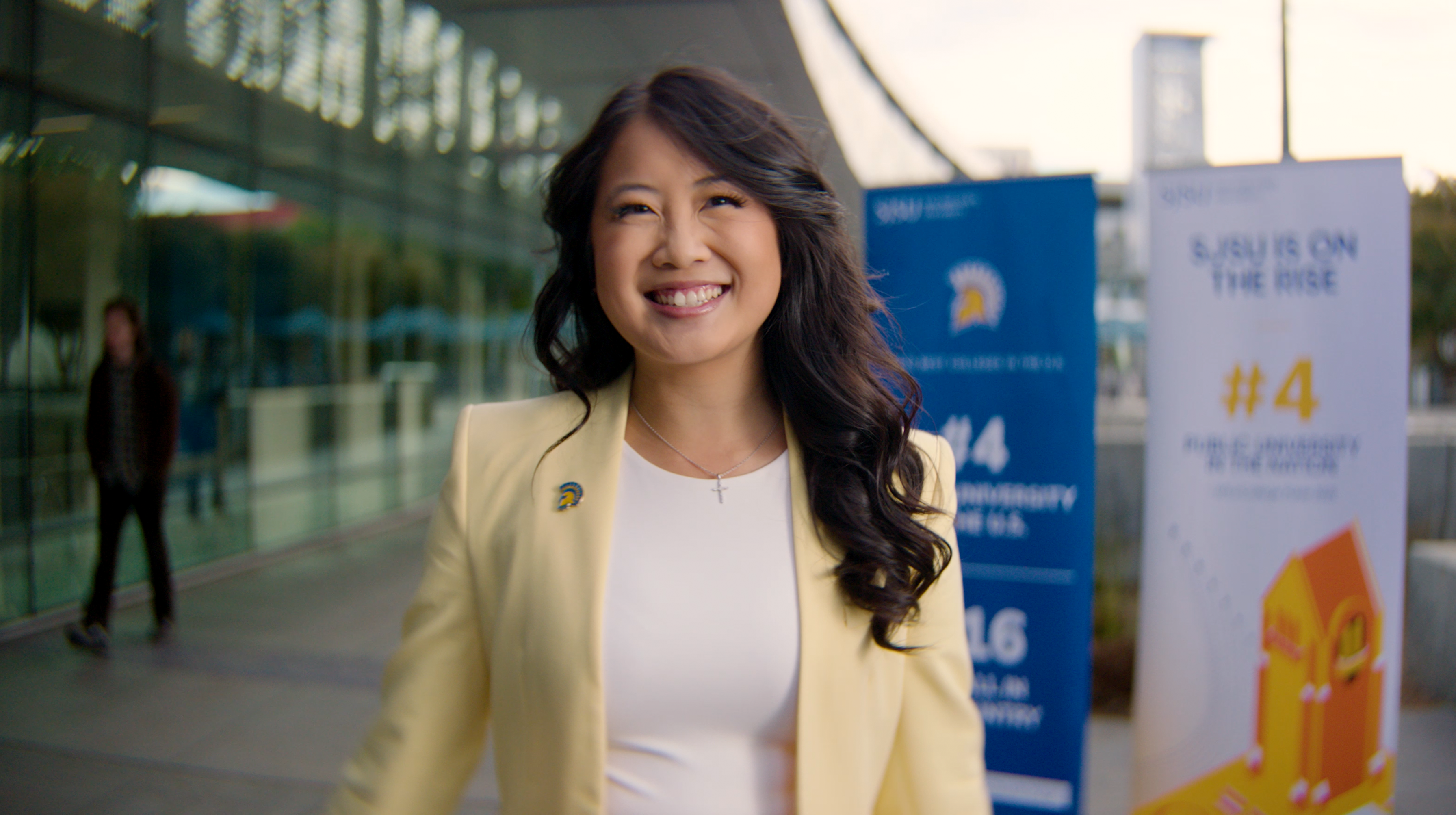 An Asian American female, Nina Chuang, wears a yellow jacket and smiles in front of two rankings banners in the SJSU On the Rise video